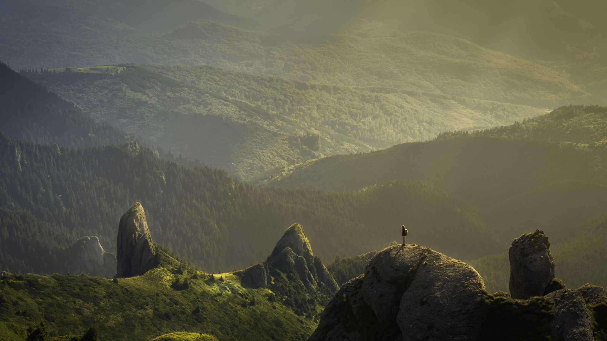 Person standing on a rocky outcrop overlooking a mountainous landscape with a warm, golden light.